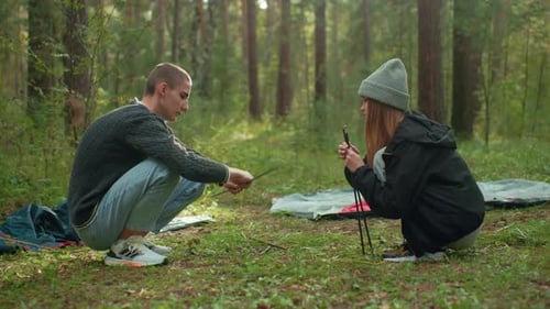 Man Fixing Tent Pole While Lady Hands Over Another in Forest Camp Setup