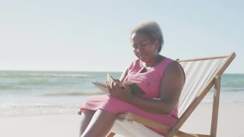 Happy senior african american woman sitting on deck chair and reading book at beach, in slow motion