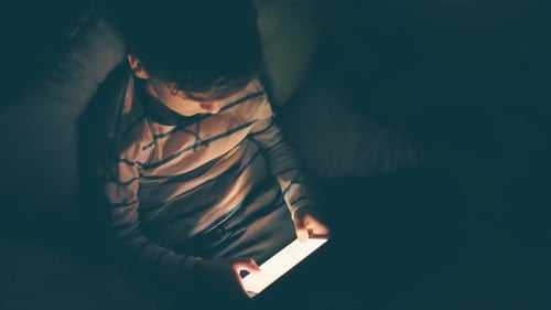 Un garçon souriant heureux est allongé dans son lit sur des coussins, des émotions, joue sur un smartphone, un jeu sombre