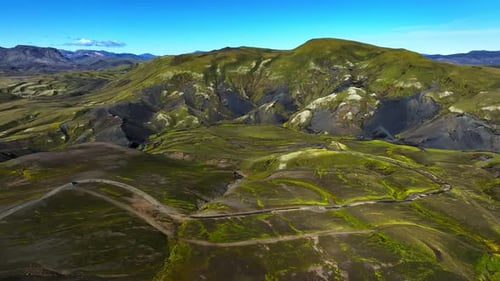Drone sweep over mossy volcanic highlands with winding dirt tracks beneath a bright blue sky,