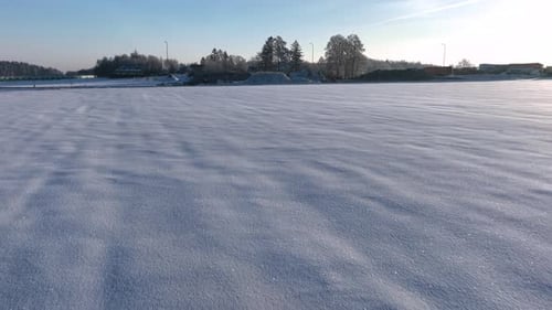 A vast snowy field under a clear blue sky, trees and buildings in the distance. Smooth panning