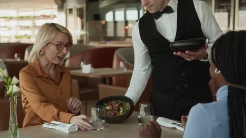 Diverse Girlfriends Chatting in Modern Restaurant while Waiter Serving Dishes