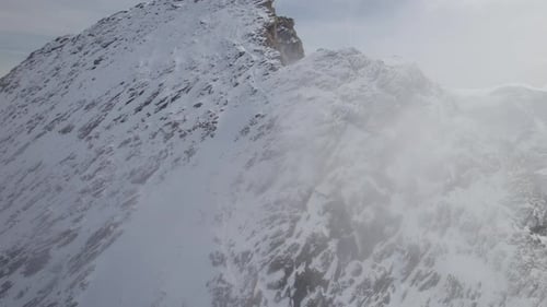 Aerial orbit shot of flight along rocky and snowy mountain wall in clouds of sky
