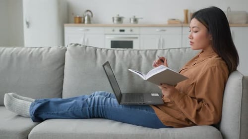 Side View Asian Woman Girl on Sofa with Laptop Student Writing Notes in Copybook Preparing for Exam
