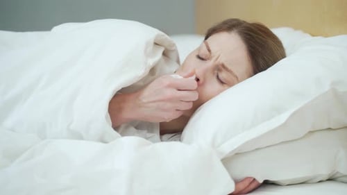 Femme blonde malade se mouchant, éternuant et toussant allongée sur le lit chaud de la chambre