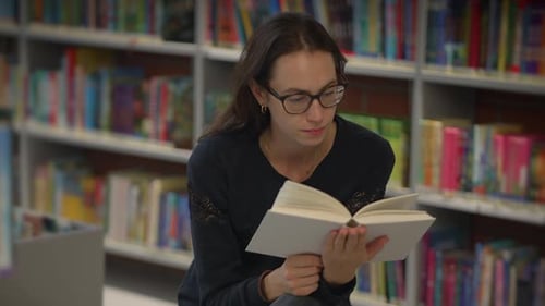Young Woman Engaged in Reading a Book in a Library Surrounded By Colorful Shelves