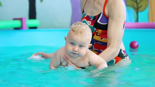 Little cute blond baby lying on woman’s hand in the swimming pool.