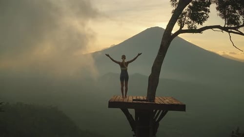 Slender woman on wooden tree platform raising arms into greeting pose, mount Agung