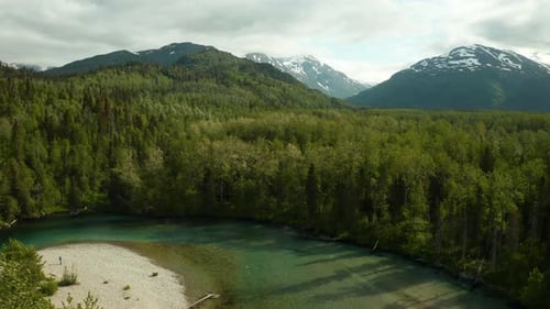 Dramatic aerial footage of green river bend with high mountain peaks rising in the background