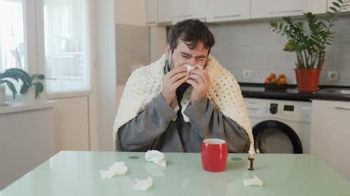 Seasonal Discomfort Scene Duvet Clad Man with Flu Shivering and Wiping Nose in Kitchen Setting