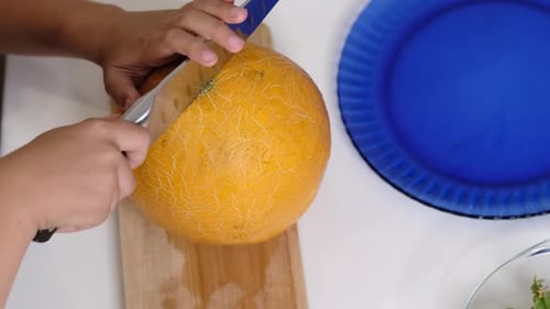 Nahaufnahme von Händen, die eine ganze Melone auf einem Holzschneidebrett schneiden, mit einem blauen Teller in der Nähe dieses Bildes