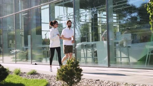 Happy Friends in Sportswear Running Along Glass Wall of Office Building in Town Young Caucasian Man