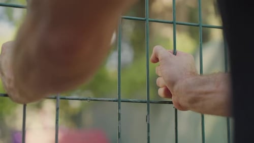 Adult hands gripping a metal wire fence, soft park or playground greenery in the background.