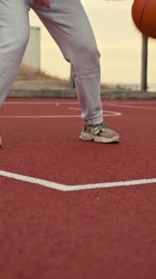 Gros plan : une fille en pantalon blanc dribble un ballon de basket orange sur un terrain de rue
