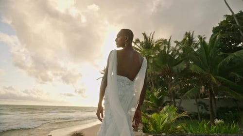 Nonbinary Black Model in Wedding Dress Poses on Tropical Beach Near Ocean at Sunset Androgynous