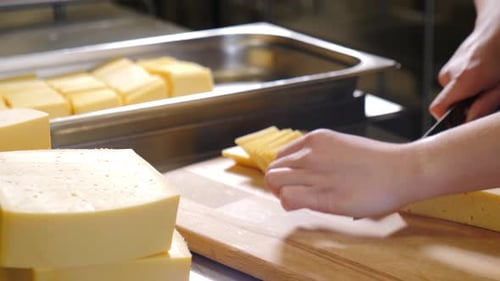 Worker is Using a Knife to Slice a Block of Pale Yellow Cheese with Small Holes on a Wooden Cutting