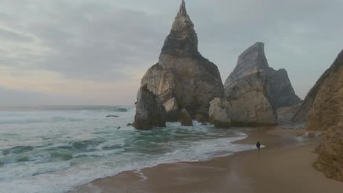 A guy running on a rocky sand beach surrounded by cliffs. Drone dolly shot. Ursa Beach, Portugal