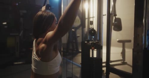 Sporty Woman Training Back Muscles on Exercise Machine in Dark Atmospheric Gym