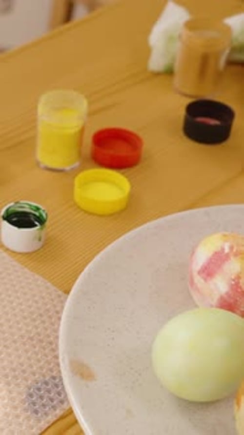 Vertical video. Woman decorating an Easter egg with green dots in a bright, sunny room.
