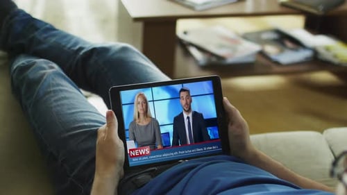 Relaxed Man Lying on His Couch Watches News Broadcast on His Tablet Computer.