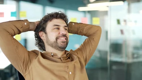 Happy businessman relaxes with hands behind head sitting at workplace in business office. Smiling
