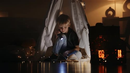 A Cute Blonde Boy Reads a Book By the Light of a Lantern Sitting at Home in a Hut in the Dark By the