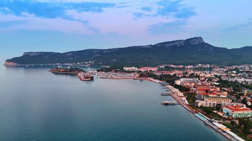 Resort town with hotels, beach line and berths. Wooded green mountain range at backdrop.
