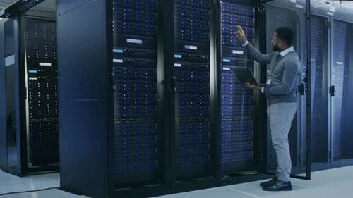 Black IT Specialist is Working on Laptop in Data Center while Standing Before Server Rack. Running