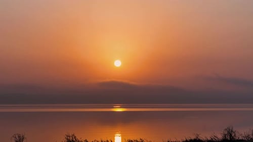 Sun rises across orange haze cloud sky above calm lake below, stunning time lapse