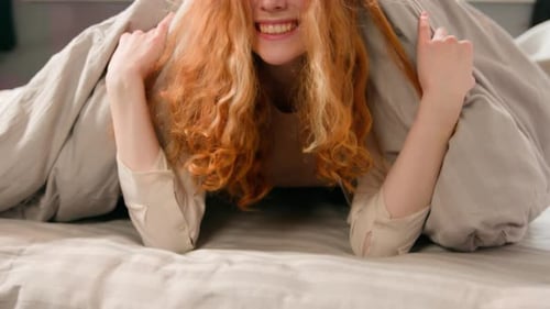 Garota feliz caucasiana sorrindo, espreita debaixo do cobertor, parece brincalhona se escondendo em um branco confortável