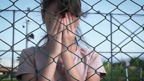 Sad Child Behind Wire Fence Covers Her Eyes