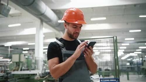 Man in uniform and hard hat stands in the factory and uses phone