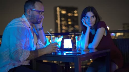 Unhappy couple sits on terrace in city bar drinking beverage at night