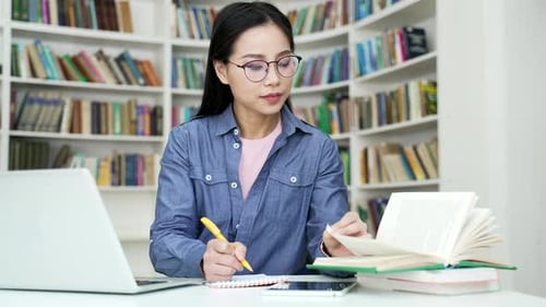 Asian female student studies by reading books, taking notes in campus library space. Young woman