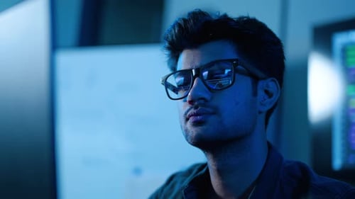 Un joven desarrollador de software o profesional de TI que lleva gafas analiza el reflejo de la pantalla de datos por la noche