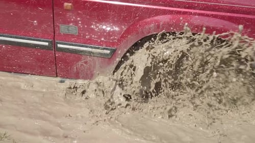 A red vehicle drives through deep muddy waters in slow motion. Close-up of the tire splashing dirty