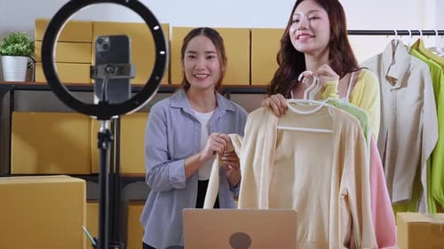 Two young asian woman live streaming online clothing store while showing clothing with smartphone.