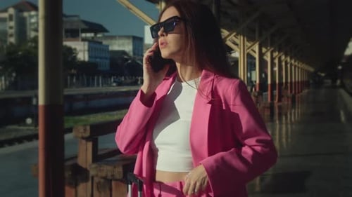 A Gorgeous Woman in a Pink Suit is Standing at the Train Station Talking on Her Mobile Phone