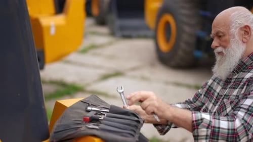 Side View Portrait of Concentrated Confident Senior Man with Wrench Checking Tractor Outdoors