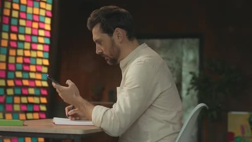 Bearded Young Man Writing in Notebook Sitting at Desk in Workplace Thinking Use Phone Typing Social