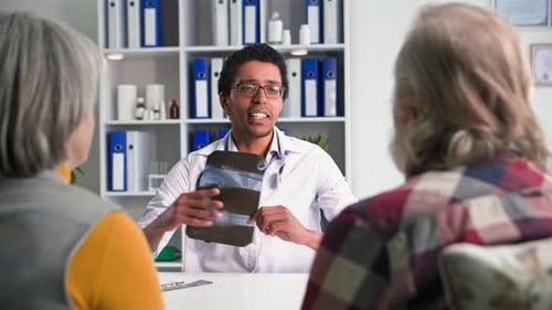 Young Black Male Doctor Communicates with an Elderly Man and a Woman Guard Holds a Xray in His Hands