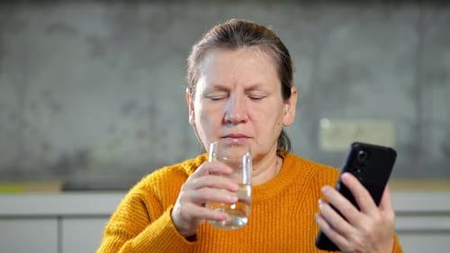 Woman Drinks Water and Looks at Phone Indoors