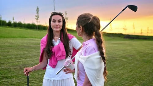 Two women dressed in white and pink clothes, holding golf clubs and talking on the course