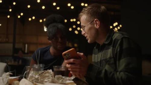 Date Of Multiracial Couple In Bar African Woman And Caucasian Man Viewing Pictures On Smartphone