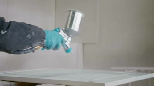 Nahaufnahme der Hände einer Schreinerin, die mit Druckluft Lack auf eine Holztischplatte sprüht.