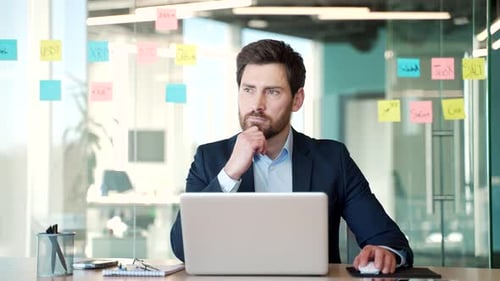 Businessman thoughtfully working in modern office with laptop. Male professional wearing formal suit