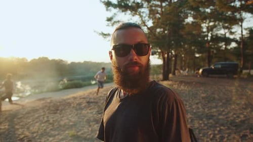 A Stylish Young Bearded Man in Glasses is Standing Next to Friends Who are Playing Ball on the River