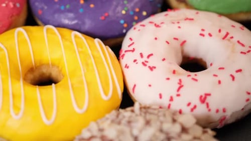 In Nahaufnahme drehen sich bunte Donuts mit cremefarbener Schokoladenglasur und Streuseln auf schwarzem Hintergrund.