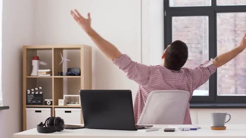 Rear view of a relaxed man in an office with stretched arms, a laptop, and a briefcase.