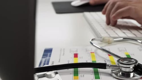 The doctor sits at the hospital table and works on his computer with a stethoscope.